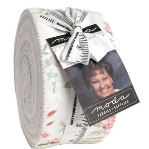 May include: A roll of fabric strips, featuring a floral pattern in soft pastel colors. The fabric is tied with a white ribbon printed with a ruler design and the words "moda FABRICS + SUPPLIES". A tag with a woman's photo and the moda logo is attached.