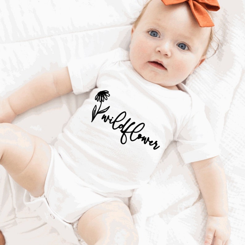 May include: White baby onesie with black text that reads "wildflower" and a small black flower graphic.