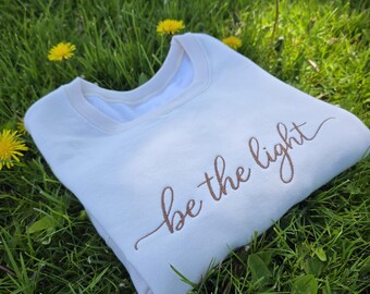 Op de afbeelding: Crèmekleurige sweatshirt met de geborduurde tekst "be the light" in sierlijke letters. Het sweatshirt ligt op groen gras met gele paardenbloemen.
