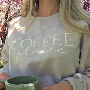 Op de afbeelding: Een lichtgrijs sweatshirt met de geborduurde tekst "COFFEE please" in crème. Een persoon houdt een groene keramische mok vast. De sweatshirt heeft lange mouwen en een ronde hals.