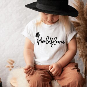 May include: A white t-shirt with a black daisy and the text "wildflower" printed on the front. A young person is wearing the shirt and a black hat.