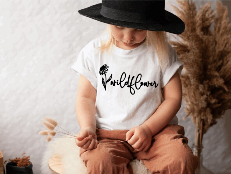 May include: A white t-shirt with a black daisy and the text "wildflower" printed on the front. A young person is wearing the shirt and a black hat.