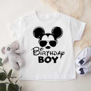 May include: White t-shirt with a black and white illustration of Mickey Mouse wearing sunglasses. The text "1st Birthday Boy" is printed below the illustration.