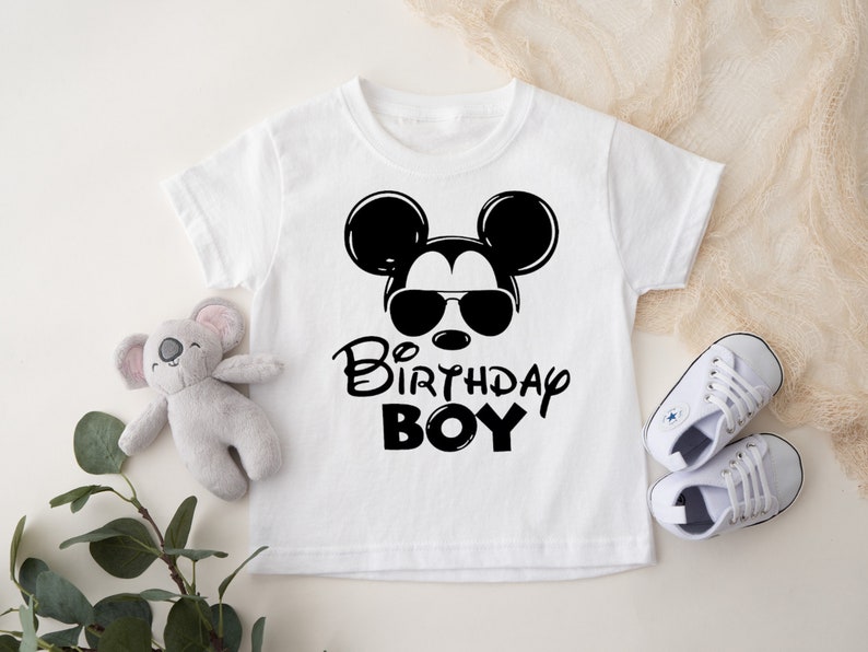 May include: White t-shirt with a black and white illustration of Mickey Mouse wearing sunglasses. The text "1st Birthday Boy" is printed below the illustration.