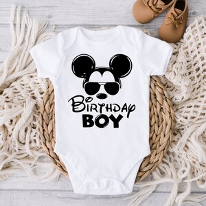 May include: White baby onesie with a black and white illustration of Mickey Mouse wearing sunglasses. The text "Birthday Boy" is printed below the illustration.