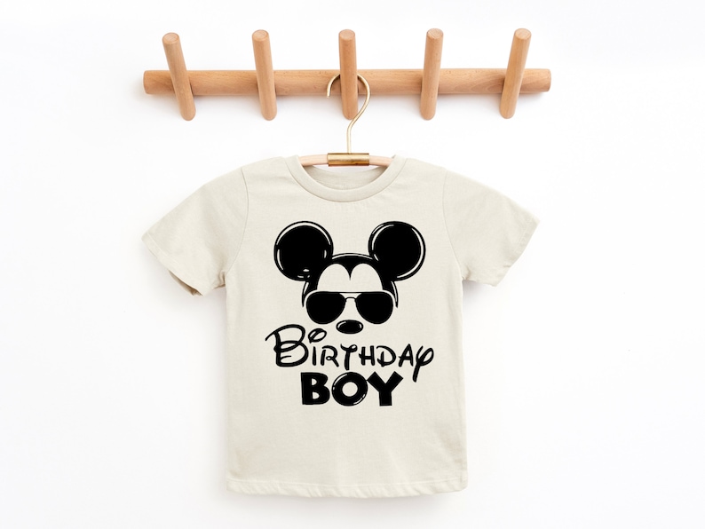 May include: A beige short-sleeved t-shirt with a black Mickey Mouse graphic wearing sunglasses and the text "Birthday Boy".