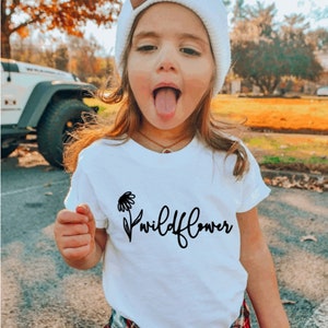May include: A young person wearing a white t-shirt with a black wildflower graphic and the text "Wildflower". They are also wearing a white knit beanie, a red and black plaid shirt tied around their waist, and blue jeans.