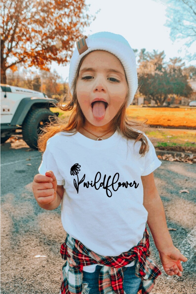 May include: A young person wearing a white t-shirt with a black wildflower graphic and the text "Wildflower". They are also wearing a white knit beanie, a red and black plaid shirt tied around their waist, and blue jeans.