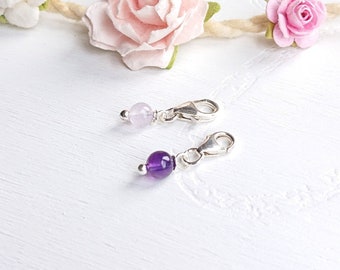May include: Two silver charm pendants with a light purple and a dark purple gemstone. The charms are on a white surface.
