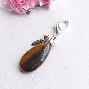 Op de afbeelding: Een druppelvormige tijgeroog edelsteen bedel met een zilverkleurige sluiting, veer, ster en initiaal "H" hanger. De gepolijste steen toont bruine en gouden banden. De bedel is waarschijnlijk voor een armband of ketting.