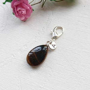 Op de afbeelding: Een druppelvormige tijgeroog-bedel met een zilveren sluiting en een kleine schijf met de letter "A". De bedel is bruin met donkerbruine strepen. Een roze roos staat op de achtergrond.