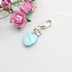 May include: A silver-colored clasp with a light blue heart-shaped charm and two small round charms with the letters "W" and "T". Pink paper roses are in the background.