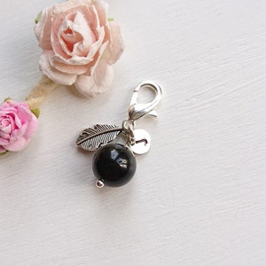 May include: A silver charm featuring a black bead, a silver feather charm, and a silver letter charm. The charm is attached to a silver lobster clasp.
