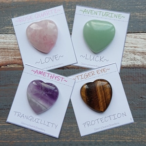 Könnte beinhalten: Vier herzförmige Kristallsteine auf weißen Karten mit Text, der ihre Eigenschaften beschreibt. Die Steine sind Rosenquarz, Aventurin, Amethyst und Tigerauge. Der Text auf den Karten lautet: ~ROSE QUARTZ~ ~LOVE~ ~AMETHYST~ ~TRANQUILLITY~ ~AVENTURINE~ ~LUCK~ ~TIGER EYE~ ~PROTECTION~