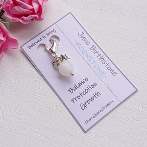 May include: A silver charm with a white moonstone bead and the letter K. The charm is attached to a silver clip. The card reads "June Birthstone ~Moonstone~ Balance Protection Growth CherryCharmJewellery"