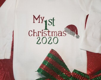 May include: White long-sleeved baby onesie with green and red glitter text that reads "My 1st Christmas 2020" and a red glitter Santa hat. A red and green striped bow is in the foreground.