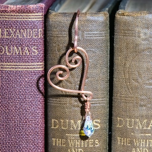 Könnte beinhalten: Ein herzförmiges Lesezeichen aus Kupferdraht mit einem klaren Kristallanhänger, der an der Unterseite hängt. Das Lesezeichen befindet sich zwischen zwei Büchern mit dem Text "ALEXANDER DUMAS" auf dem Rücken.