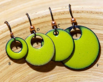 Pode incluir: Quatro brincos circulares verde-limão com bordas escuras e recortes centrais, suspensos por ganchos de metal escuro com pequenas contas cor de cobre. Os brincos estão marcados com as letras B, D, C e F para indicar os tamanhos.