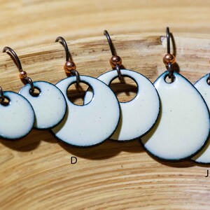 Pode incluir: Cinco pares de brincos de esmalte branco com contornos pretos. Os brincos são de diferentes formas, incluindo círculos e ovais. Os brincos estão pendurados em uma superfície de madeira.