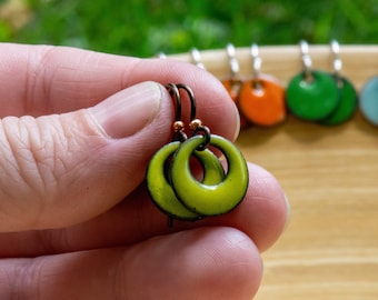 May include: A pair of lime green circular enamel earrings with a glossy finish and dark metal edges. The earrings feature a small round cutout in the center and are attached to dark metal ear wires with a small copper-colored bead accent.
