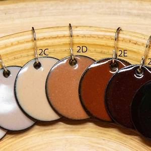 May include: A collection of seven circular enamel pendants labeled 2A through 2G, featuring a gradient of colors from white to black. Each pendant has a silver-toned metal hook and a smooth, glossy finish, arranged in a row on a curved surface.