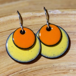 May include: A pair of earrings with two layered circles. The top circle is orange with a black outline and the bottom circle is yellow with a black outline and a light blue edge.