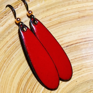 May include: A pair of long, teardrop-shaped earrings featuring a glossy, vibrant red enamel finish on the front surface. The earrings are suspended from dark metal hooks, each accented with a small, polished copper-colored bead at the top.