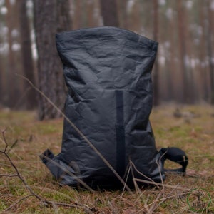 May include: A black, crumpled, waterproof backpack with adjustable straps. The backpack is sitting on a bed of moss and pine needles in a forest setting.