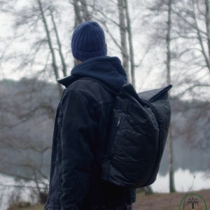 May include: A person wearing a blue beanie and a black denim jacket with a large black backpack. The person is standing in a forest with trees and a lake in the background.
