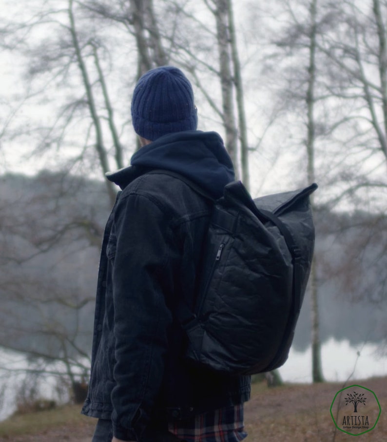 May include: A person wearing a blue beanie and a black denim jacket with a large black backpack. The person is standing in a forest with trees and a lake in the background.