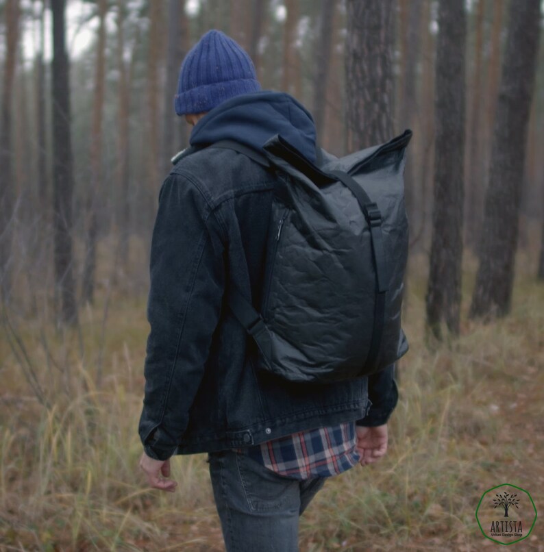 May include: A person wearing a blue beanie, a black denim jacket, a plaid shirt, and blue jeans is walking through a forest. They are carrying a black backpack with a roll-top closure.