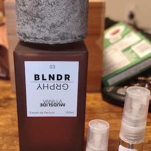 May include: A brown perfume bottle with a textured gray cap, labeled "BLNDR GRPHY VANILLA MUDSLIDE." Includes two smaller sample vials, one labeled 2 ML and the other 5 ML. The main bottle contains 100ml of perfume.