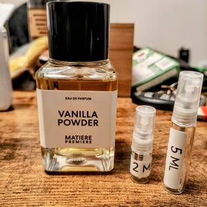 May include: A perfume bottle with a black cap and a white label that reads "Vanilla Powder" and "Matiere Premiere." Two smaller sample vials are next to the bottle, one labeled "2 M" and the other "5 ML."