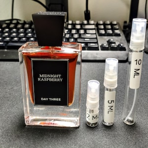 May include: A clear glass perfume bottle with a black cap and the words "MIDNIGHT RASPBERRY" and "DAY THREE" printed on the front. There are three small, clear spray bottles labeled 2 mL, 5 mL, and 10 mL.