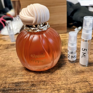 May include: A light orange perfume bottle with a cream-coloured decorative top and a gold-coloured band. The bottle is labelled "Vanilla Voyage" and includes two small sample spray bottles, one labelled "2 ML" and the other "5 ML".