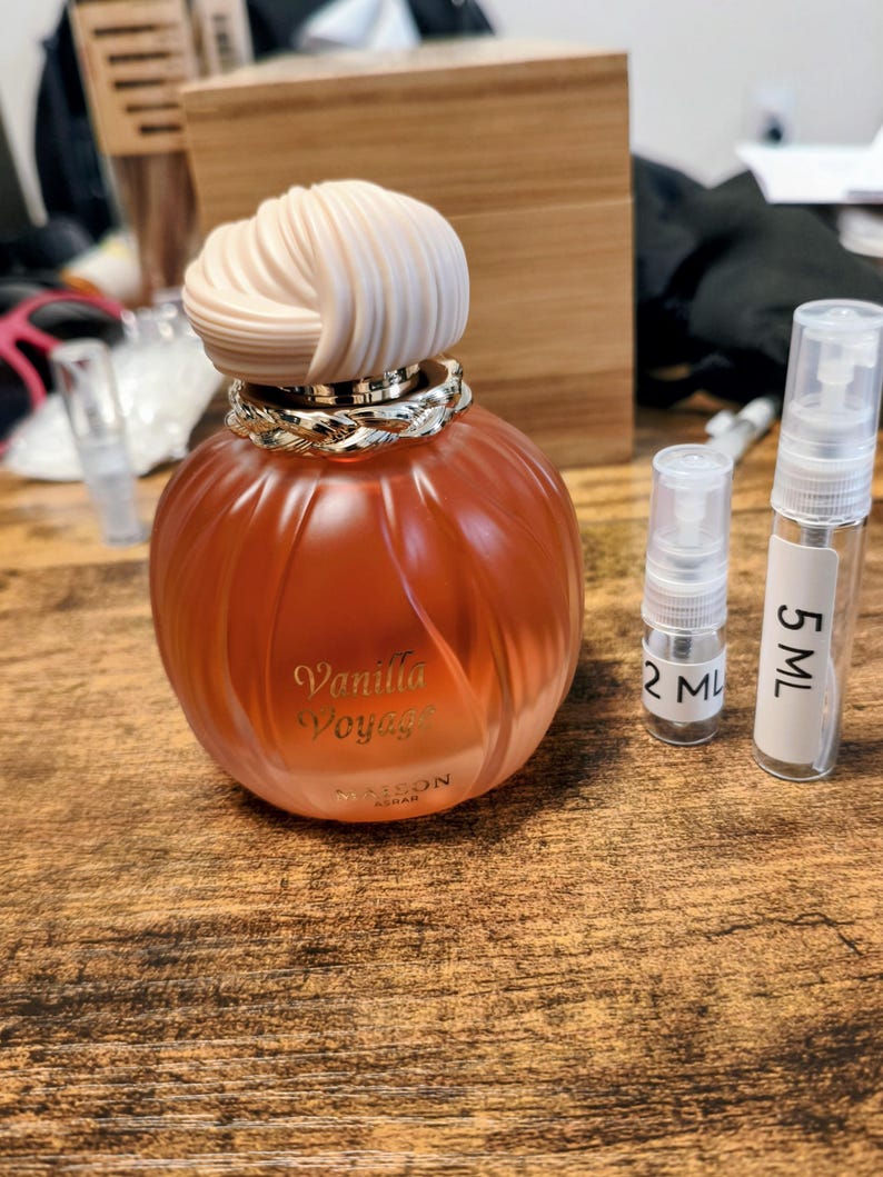 May include: A light orange perfume bottle with a cream-coloured decorative top and a gold-coloured band. The bottle is labelled "Vanilla Voyage" and includes two small sample spray bottles, one labelled "2 ML" and the other "5 ML".