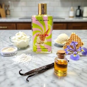 May include: A perfume bottle with a gold cap and a label that says "Lick Me" with an ice cream cone design. The image includes vanilla beans, whipped cream, and a purple iris flower, suggesting a dessert-inspired fragrance.