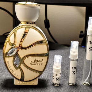 May include: A perfume bottle with a cream and gold design, the text "Vanille Blanc" and "Sawaar" is visible. There are three small sample bottles labeled 2 ML, 5 ML, and 10 ML. The perfume bottle has a decorative jewel.