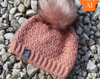 May include: A dusty rose crochet beanie featuring a ribbed brim and a large, fluffy faux fur pom-pom on top. A small, rectangular grey leather-like tag is attached to the side of the brim, embossed with a mountain graphic and the letters CMD.