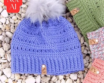 May include: A periwinkle blue crocheted beanie featuring a large, fluffy white faux fur pom-pom on top. The hat has a ribbed brim and a small, rectangular tan leather tag with the letters CMD and a mountain graphic attached to the front.