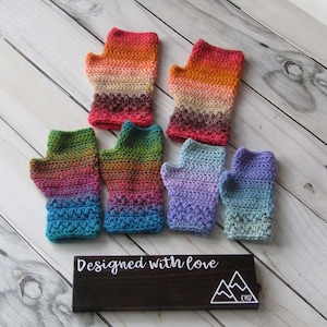 Pode incluir: Seis pares de luvas sem dedos de crochê coloridas, com um degradê de cores, sobre um fundo de madeira branca. As luvas estão dispostas em duas filas de três. O texto "Designed with love" está em uma placa preta abaixo das luvas.