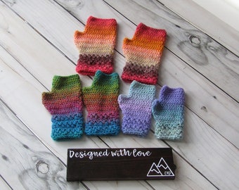 May include: Six pairs of colorful crocheted fingerless gloves, with a gradient of colors, on a white wood background. The gloves are arranged in two rows of three. The text "Designed with love" is on a black sign below the gloves.