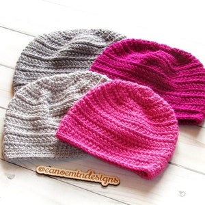 May include: Three crocheted hats, one gray and two pink, are stacked on a white wooden surface. The hats are made with a textured stitch and have a simple, classic design. The text "@canoemtn designs" is visible on a wooden tag.