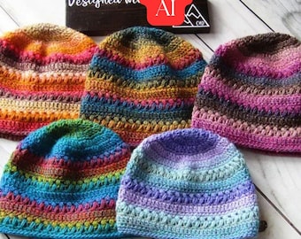 May include: Five crocheted beanies in various multi-colored yarn patterns. The hats feature horizontal stripes of orange, yellow, pink, blue, purple, and green. A dark wooden sign behind the hats displays the text Designed With Not AI.