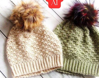 May include: Two crocheted beanies with ribbed brims and textured stitch patterns. One beanie is light beige with a brown faux fur pom-pom, and the other is olive green with a multi-coloured dark faux fur pom-pom. A red tag reads Not AI.