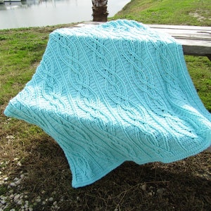 May include: A light blue crocheted blanket with a diamond pattern, draped over a wooden picnic table. The blanket is on a grassy surface with a body of water in the background.