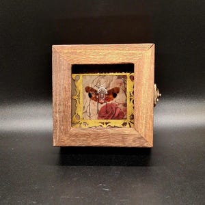 May include: A small, square wooden box with a decorative butterfly and floral design. The box features a brown wooden frame, a gold-colored border, and a vintage-style image of a butterfly and roses. The box has a small metal clasp.