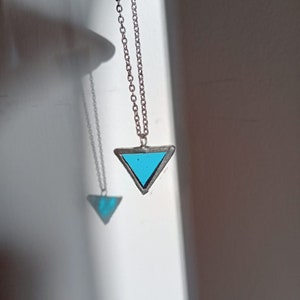 Puede incluir: Un collar de cadena de plata con un colgante triangular. El colgante está hecho de metal y tiene un centro de vidrio azul.