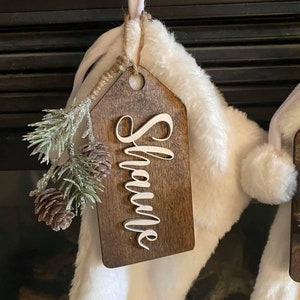 May include: A white faux fur stocking with a wooden tag that says "Shawna". The tag is brown with a white, cursive font. The stocking is decorated with a small, green and silver pine sprig with brown pine cones.