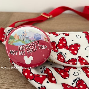 May include: A round button pin with a red background featuring a graphic of a theme park castle and a geodesic dome. The text reads DEFINITELY NOT MY FIRST TRIP with a white Mickey Mouse head icon. It is attached to a white fanny pack with red bows.
